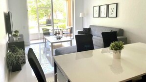 A modern living room with a sofa, a dining table, and chairs. There is a TV mounted on the wall and a large window providing natural light.