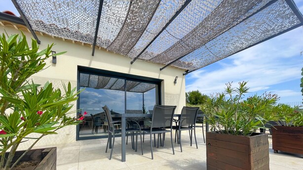 En udendørs terrasse med et bord og stole, omgivet af krukker med planter og en pergola.