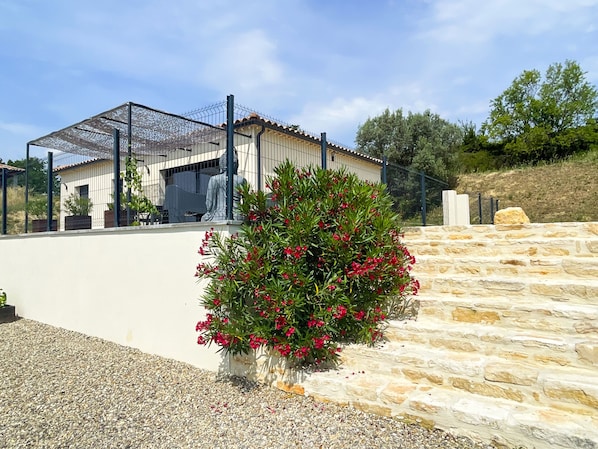 A building with a metal fence, a stone wall, and a gravel area with red flowers. - 4* villa with 2 bedrooms, air conditioning and private pool (Vagnas)