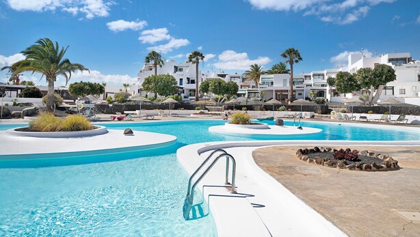 A swimming pool with a palm tree, lounge chairs, and white buildings in the background. - Studio Calimar Los Molinos with Pool, Wi-Fi & Terrace (Costa Teguise)
