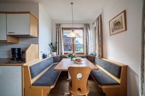 Un comedor con mesa de madera y bancos, una cafetera y un ventanal con cortinas.