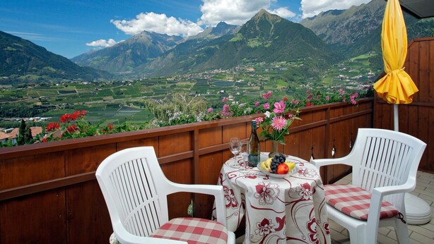 Un balcon avec des chaises en plastique blanches, une table recouverte d’une nappe à motifs floraux, une bouteille et une assiette de fruits.