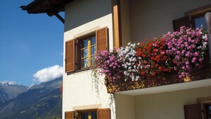 Un bâtiment doté de volets en bois, un balcon orné de plantes en suspension et un paysage montagneux en arrière-plan.