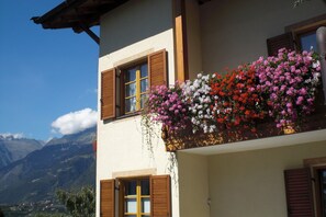 A building with wooden shutters, a balcony with hanging flowers, and a mountainous backdrop. - Apartment "Hahnenkamm Rainegg" with Mountain View, Wi-Fi, Garden & Balcony (Schenna)