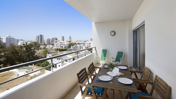 A balcony with a table set for two, chairs, a potted plant, and a view of the cityscape.