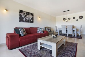 Una sala de estar con un sofá rojo, una mesa de centro de madera, un tapete estampado, una zona de comedor con mesa y sillas, y lámparas fijadas a la pared.