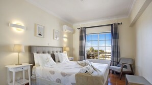 A bedroom with a bed, bedside tables, a chair, a window with curtains, and wall-mounted lights. - Estrela do Vau Apartment with Sea View, Pool, A/C, Wi-Fi, Garden, Terrace and Balcony (Portimão)