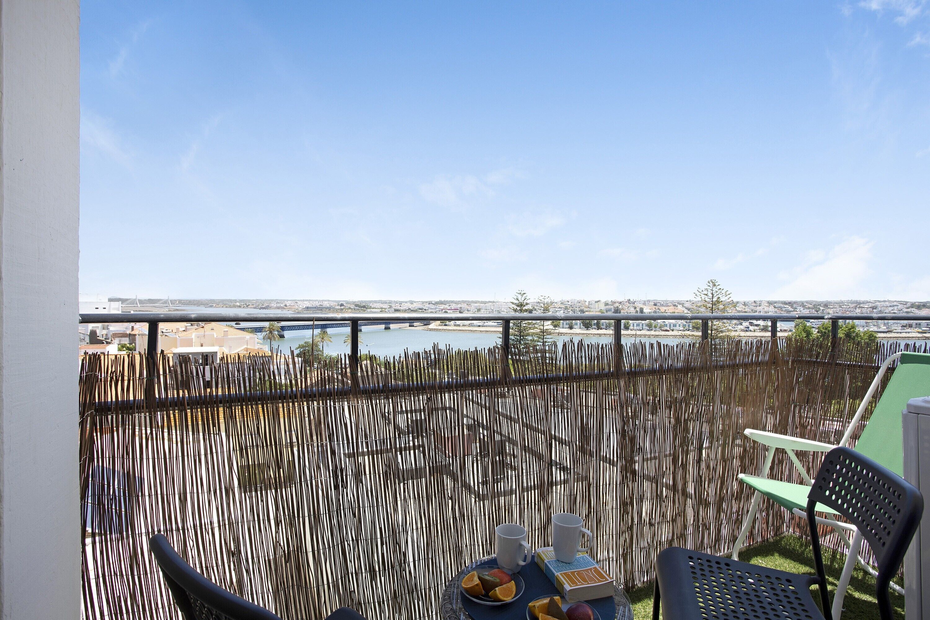 Een balkon met uitzicht op het water, een tafel met twee kopjes en een schaal fruit, en een stoel.