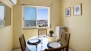 Uma área de refeições com uma mesa posta para dois, uma planta em vaso e uma janela com vista para um corpo de água e edifícios.
