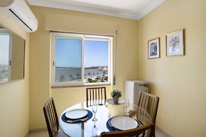 Comedor con mesa preparada para dos, una planta en maceta y ventana con vista a un cuerpo de agua y edificios.