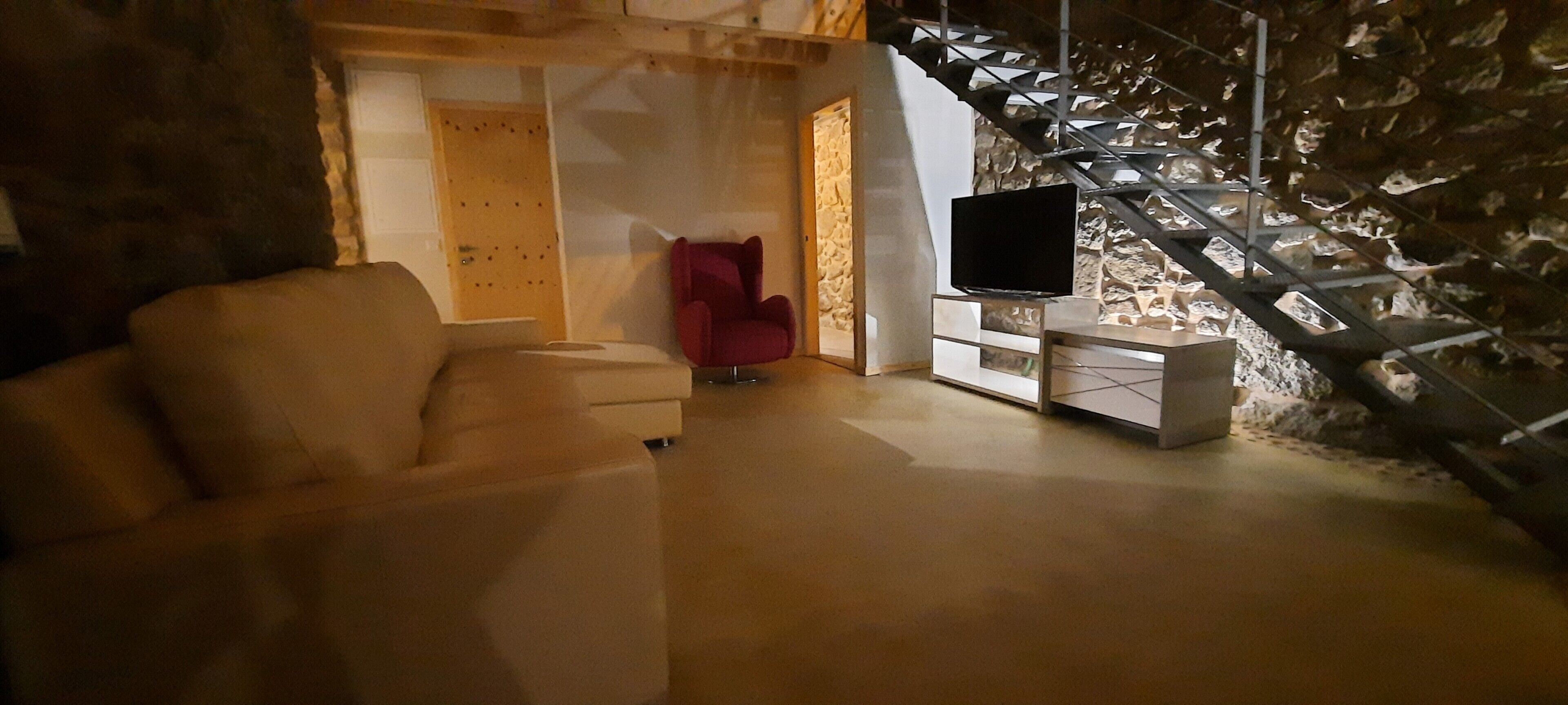 Une pièce avec un canapé, un fauteuil rouge, un téléviseur posé sur un meuble et un mur en pierre.