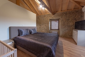 Un dormitorio con pared de piedra, techo de madera, cama con cabecera, ventana pequeña y televisor.