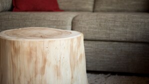 Un taburete de madera redondo que muestra un patrón de anillos de árbol, colocado frente a un sofá con un cojín rojo.