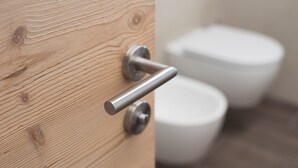 En dörr av trä med ett modernt handtag, ett synligt lås och en vit toalett i bakgrunden.