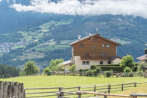 Une maison en bois dotée d’un balcon, entourée d’une cour clôturée et située dans un paysage montagneux.
