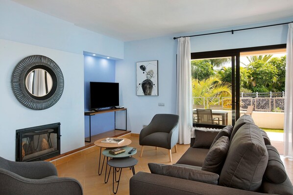 Una sala de estar moderna con chimenea, televisor de pantalla plana, sofá, sillón y comedor con vista a zonas verdes.