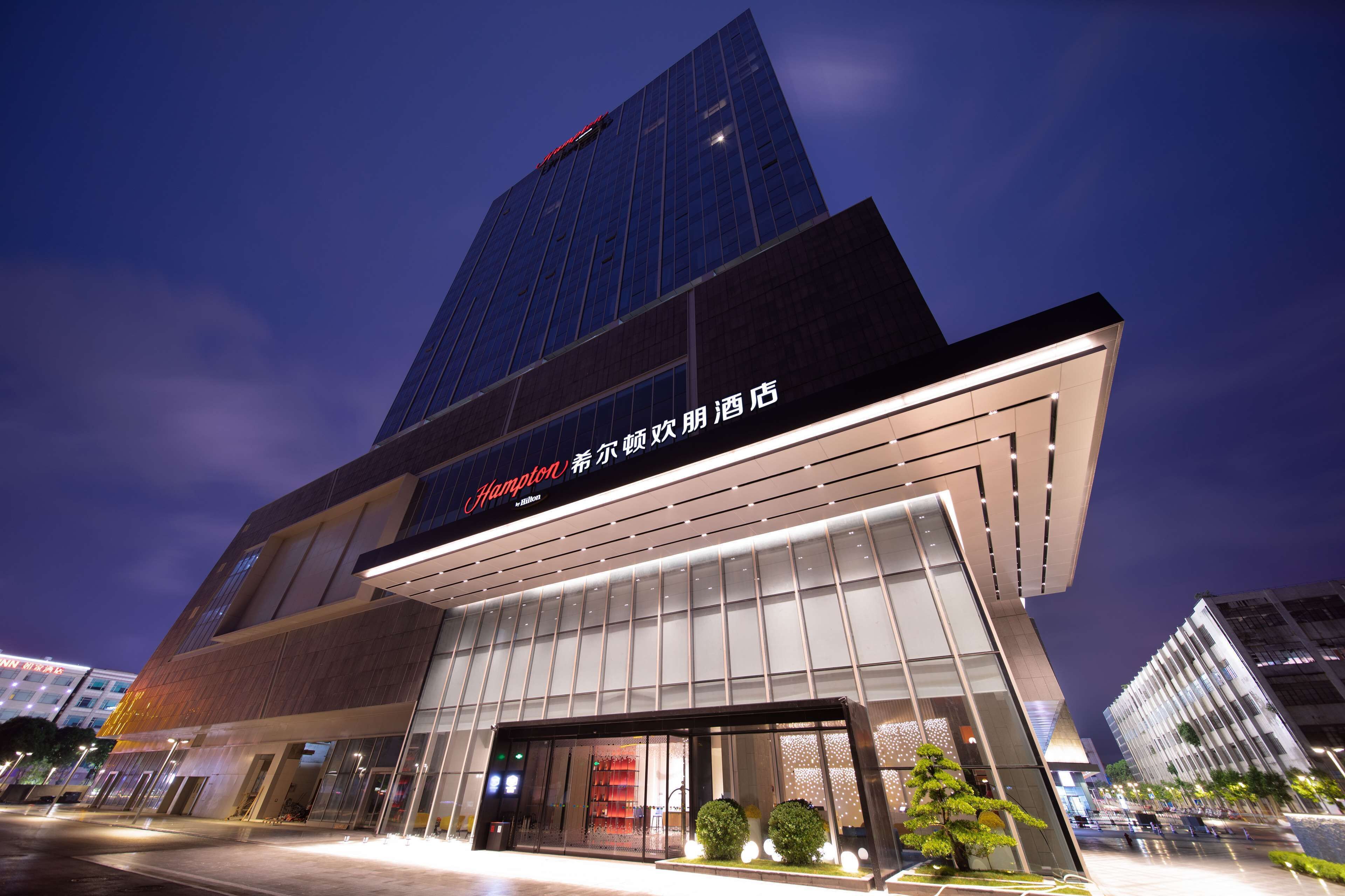 Um edifício moderno de hotel com fachada de vidro destacada e sinalização com 'Hampton' e 'Hampton by Hilton'.