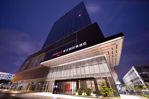 A modern hotel building with a prominent glass facade and signage for 'Hampton' and 'Hampton by Hilton'.