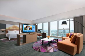 Et moderne hotelværelse med stort tv, sofa, skrivebord og byudsigt.