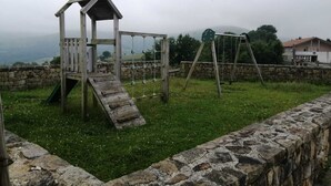 A wooden play structure with a slide and swings, surrounded by stone walls and grassy area. - Piso Ideal Para Pasar Unos Días Disfrutando de la Villa Pasiega (San Pedro del Romeral)