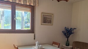 A dining area with a table set for four, a window with curtains, a framed picture on the wall, and a sofa with a vase of flowers. - Piso Ideal Para Pasar Unos Días Disfrutando de la Villa Pasiega (San Pedro del Romeral)