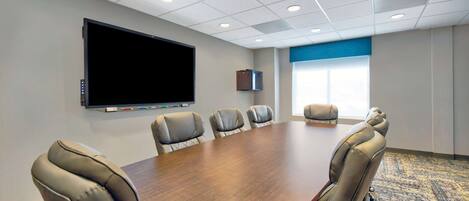 A conference room with a large table, six leather chairs, a flat-screen TV, a projector, and a window with blinds.