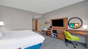 A hotel room with a large bed, a desk with a television, a mirror, and a bright yellow chair.