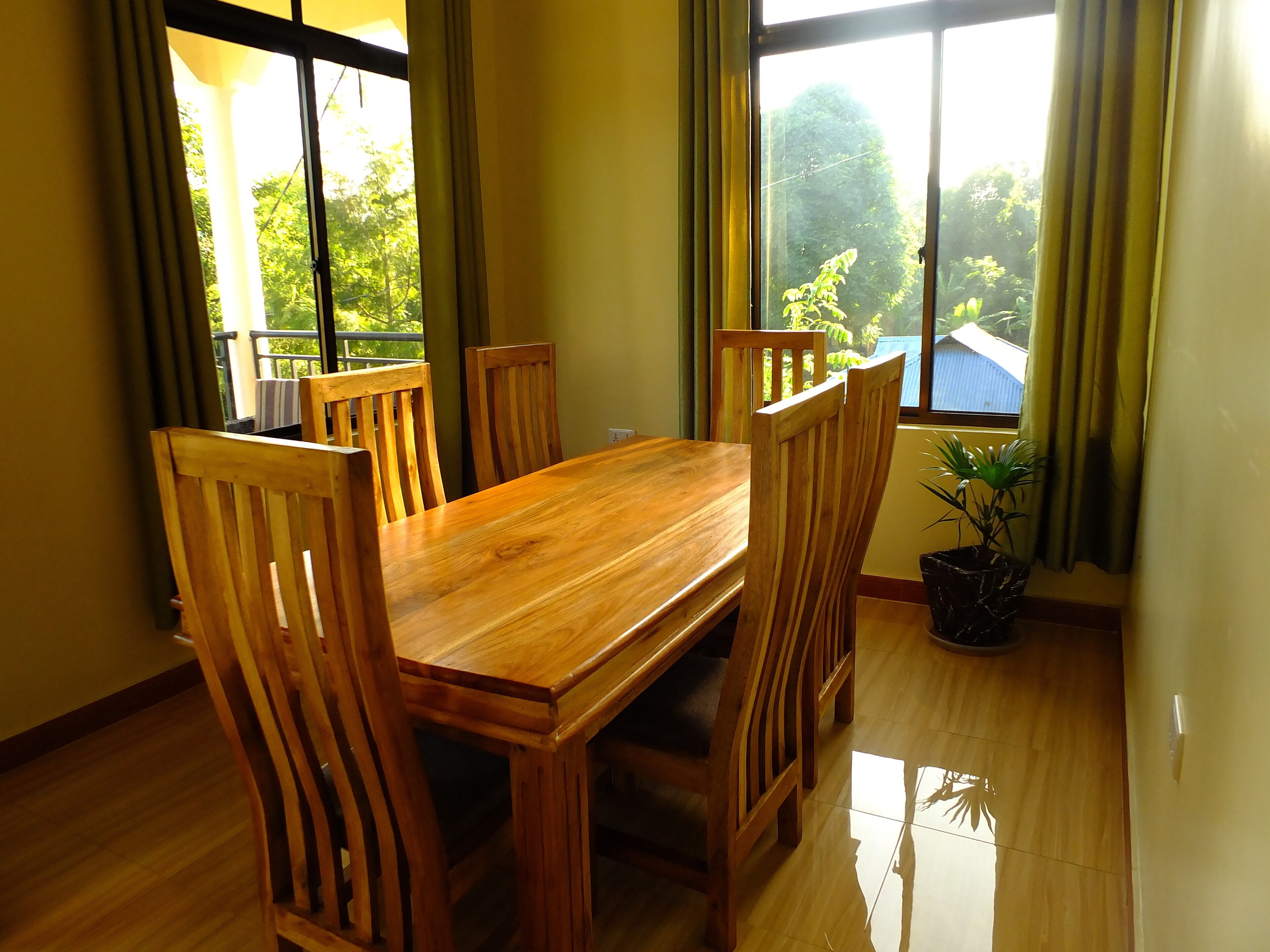 Un coin repas avec une table et des chaises en bois, de grandes fenêtres avec des rideaux et une plante en pot.