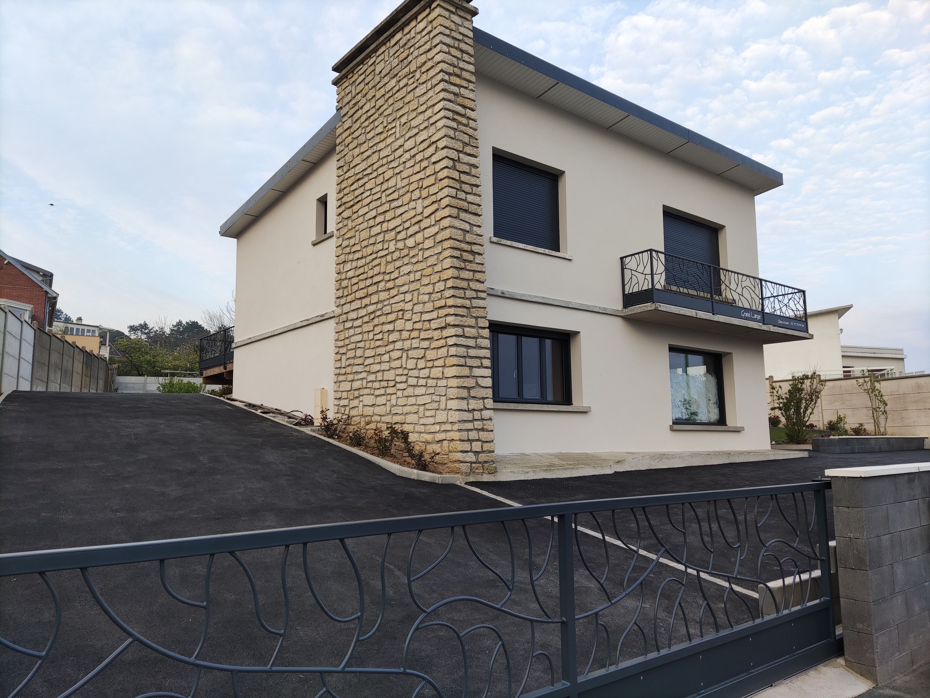 A modern two-story house with a stone facade, a balcony with metal railings, and a gated driveway.