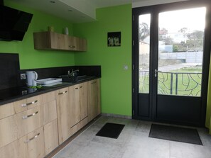 A kitchen with a black countertop, wooden cabinets, a sink, and a door leading to a balcony with a view of a residential area. - Gîte Grand Large (Petit-Caux)