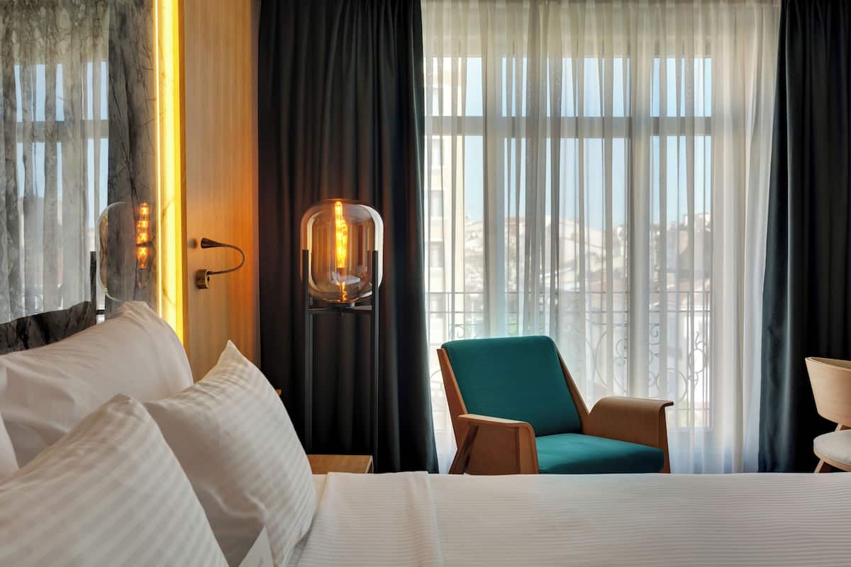 A hotel room with a large bed, a teal armchair, a wooden nightstand, and a wall-mounted lamp.
