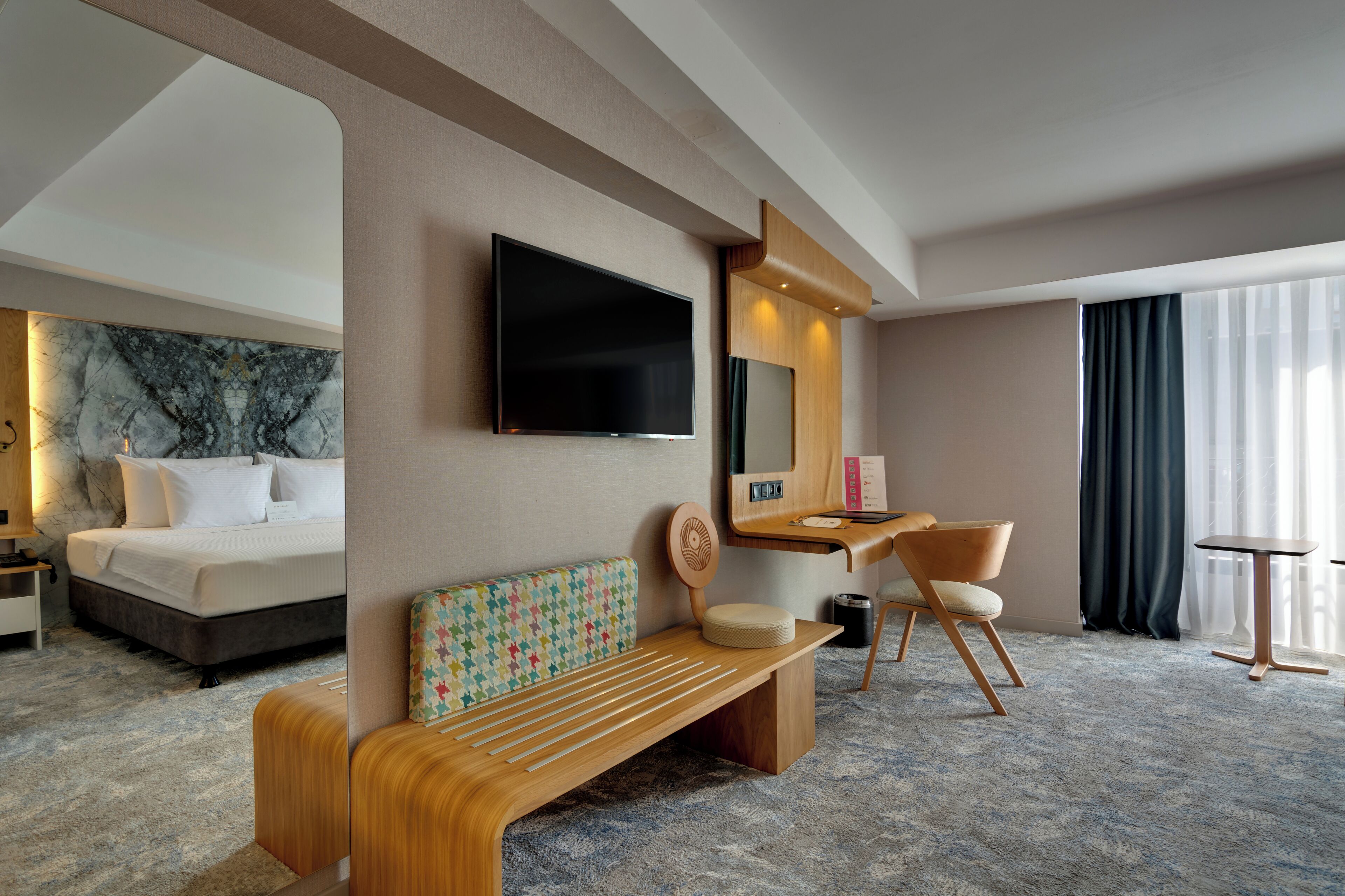 A hotel room with a bed, a desk, a chair, a bench, a TV, and a large window with curtains.
