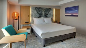 A modern hotel room with a large bed, a desk, a chair, a nightstand, a lamp, and a painting on the wall.