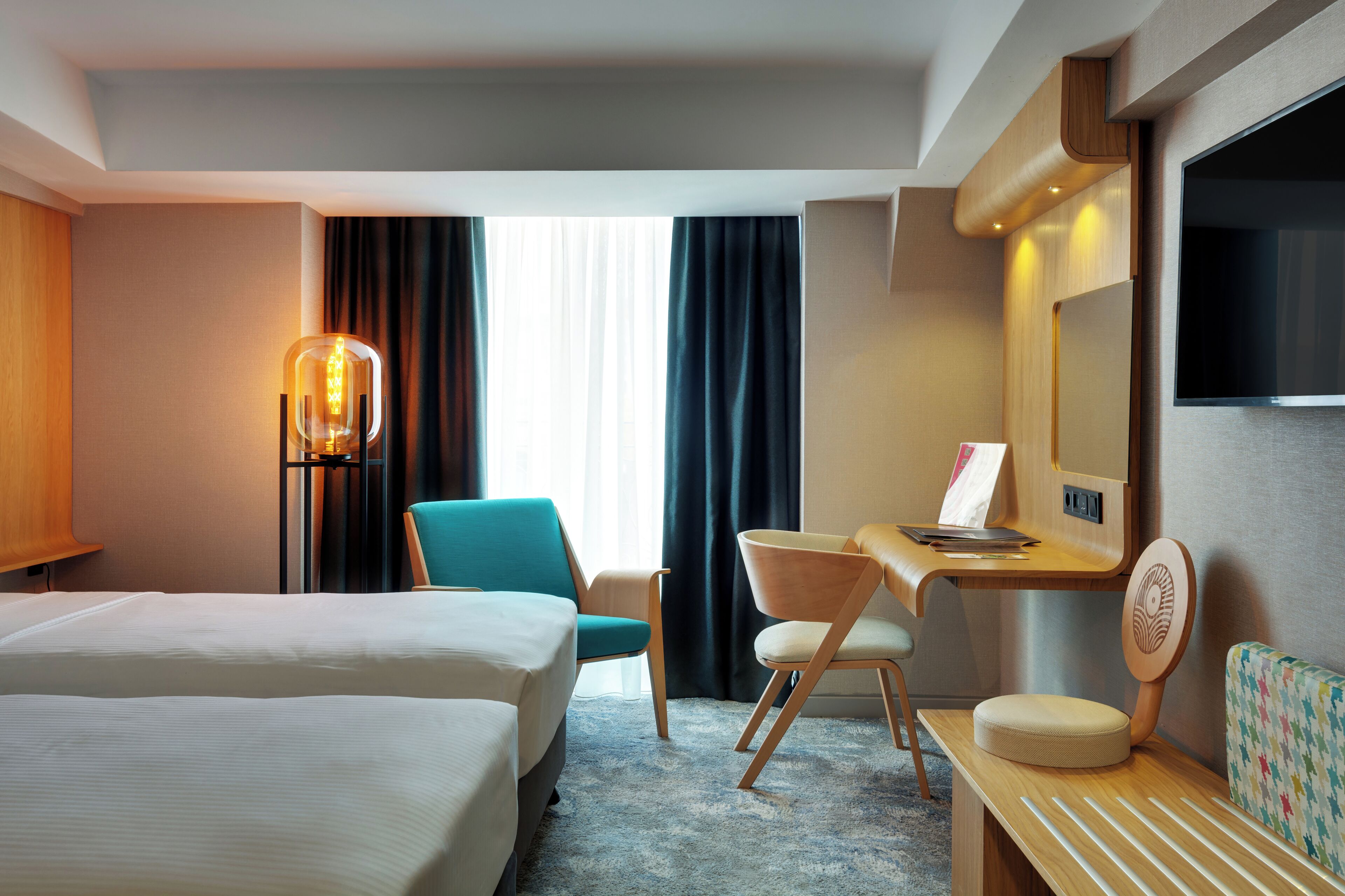 Habitación de hotel con dos camas, un escritorio, una silla, un televisor y un ventanal con cortinas.