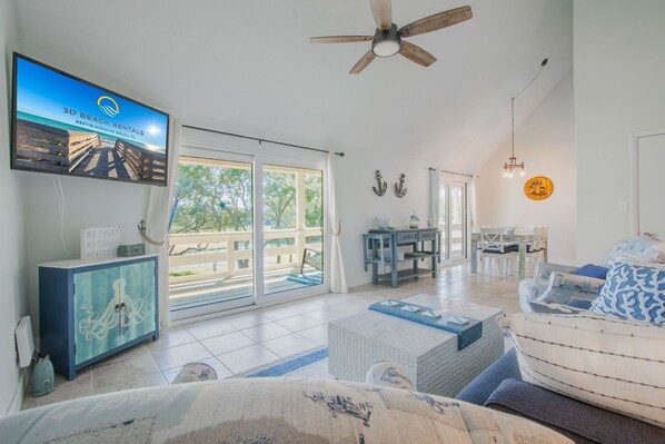 A spacious living room with a large TV displaying a beach rental ad, a ceiling fan, a dining area with a table and chairs, and a view through the glass doors.