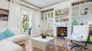 Una sala de estar con un sofá blanco, una mesa de centro, un sillón blanco y una chimenea con repisa. Se ven estanterías y un cuadro en la pared.