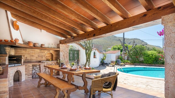Un patio exterior con chimenea de ladrillo, mesa y sillas de madera, con vistas a una piscina y un jardín.