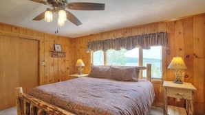 A wooden cabin bedroom with a bed, two bedside lamps, a ceiling fan, and a window with a view of the outdoors.