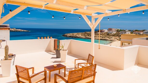 A terrace with wooden furniture, potted plants, and a view of the sea and buildings.