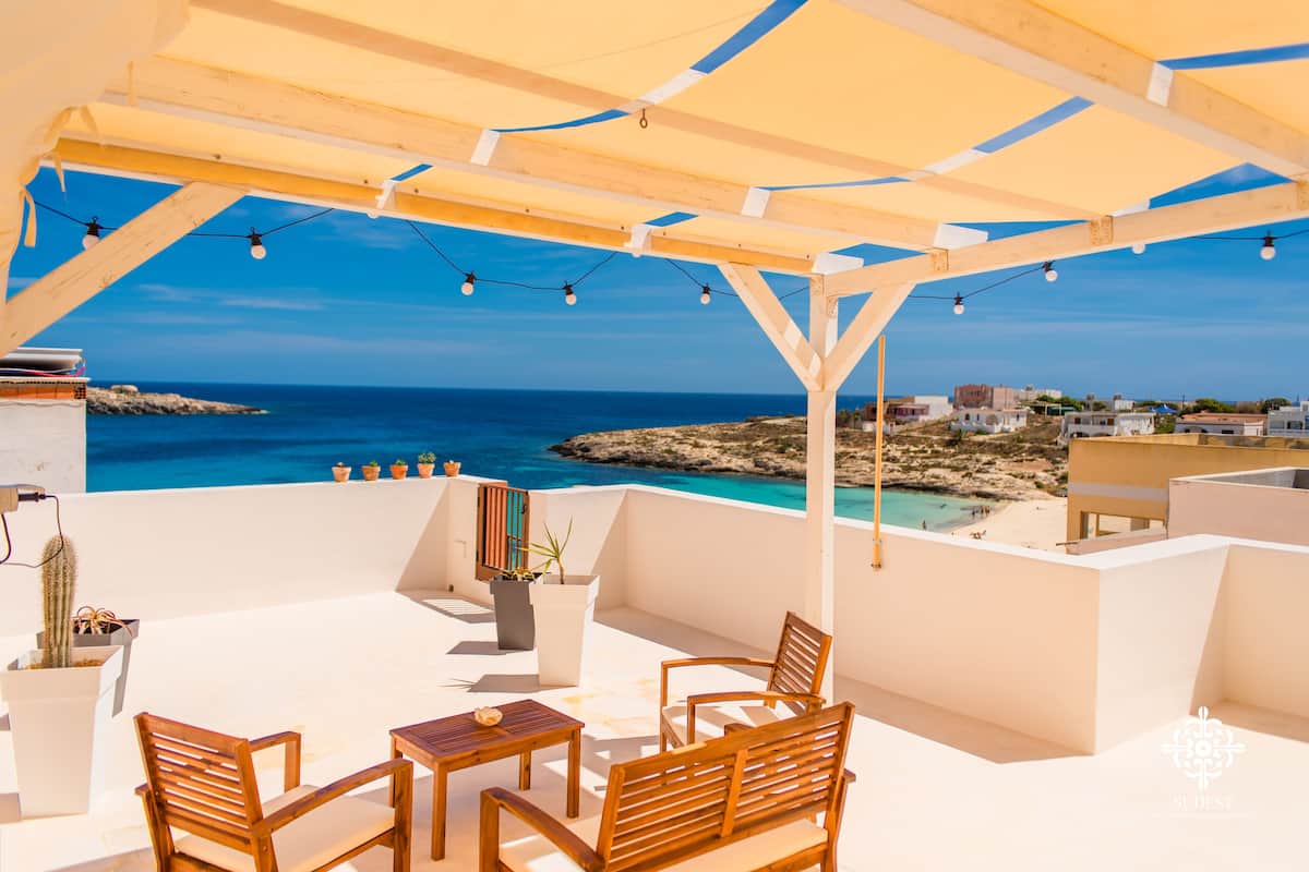 Una terraza con mobiliario de madera, plantas en macetas y vistas al mar y edificios.