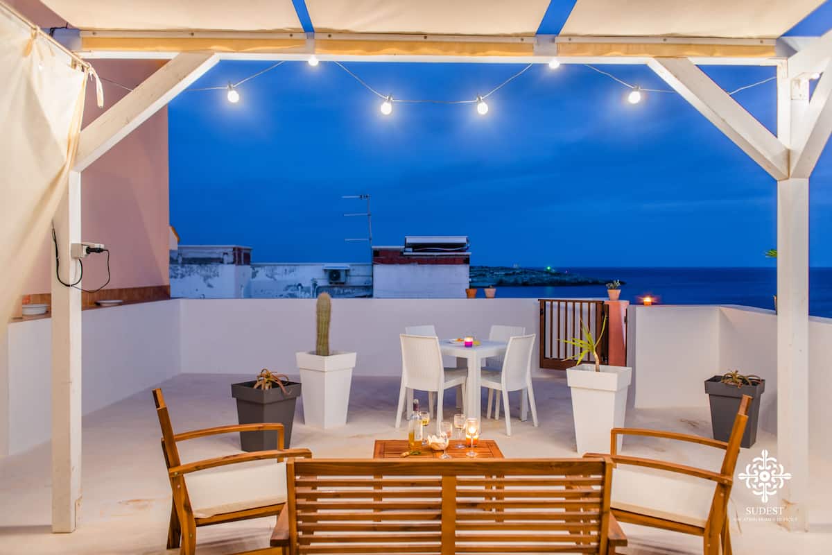 Un patio al aire libre con mobiliario blanco, luces de adorno y vista al mar al atardecer.