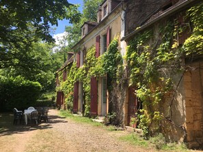 Un bâtiment rustique aux murs recouverts de lierre, avec un chemin de terre menant à une table et des chaises blanches.