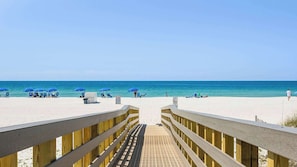 A wooden boardwalk leading to a beach with blue umbrellas and lounge chairs.