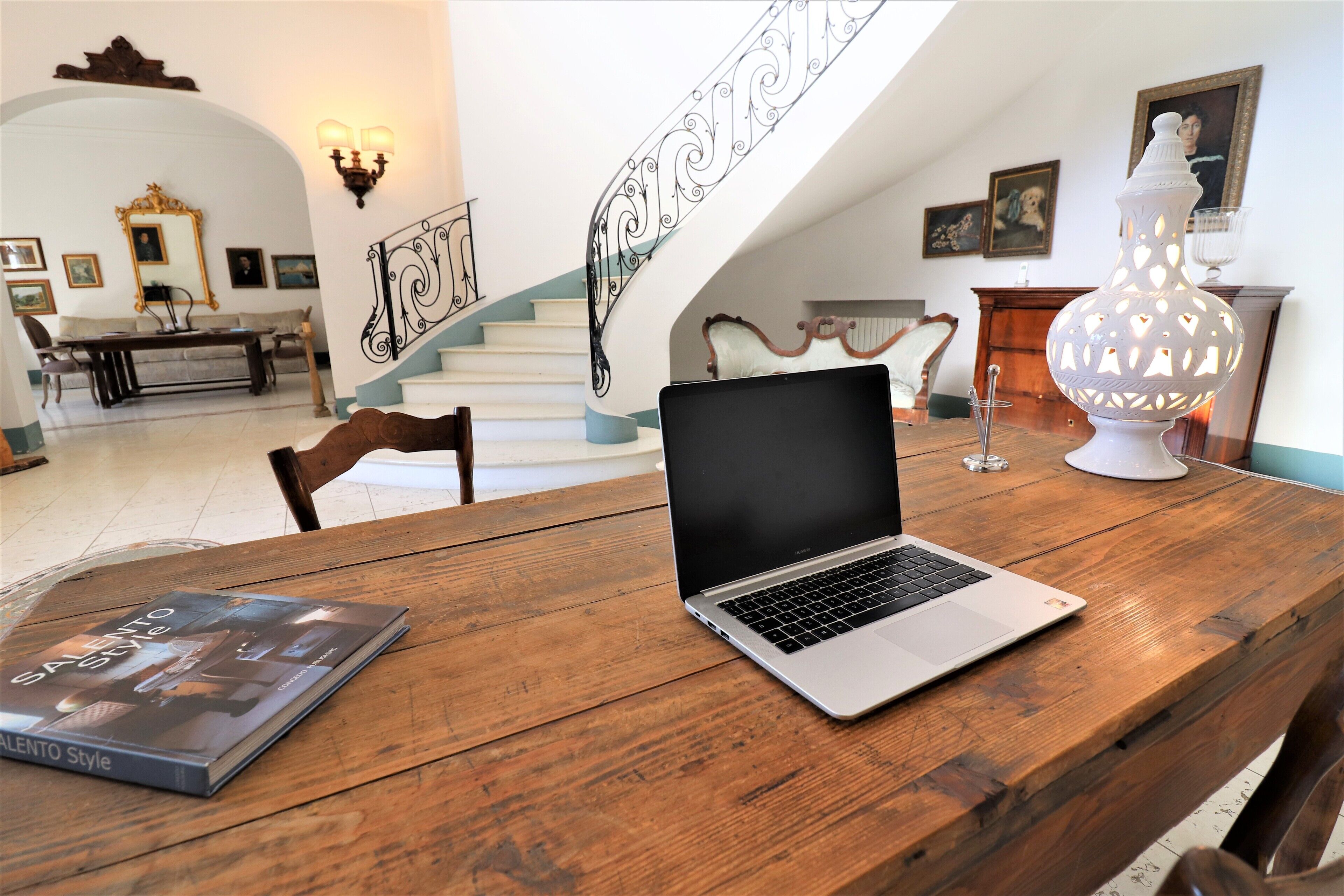 Une table en bois avec un ordinateur portable et un livre, un escalier avec des rampes décoratives et une cheminée agrémentée d’un vase ornemental.