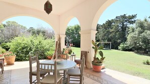 Een overdekt terras met een ronde tafel en stoelen, omgeven door potplanten en uitzicht op een gazon.