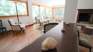 Een ruime woonkamer met een lange houten tafel, twee fauteuils en een bank. Er is een open haard en grote ramen die zorgen voor natuurlijk licht.