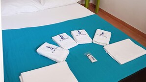 Een netjes opgemaakt bed met witte lakens, een blauwe spreideken en meerdere gevouwen handdoeken met een logo.