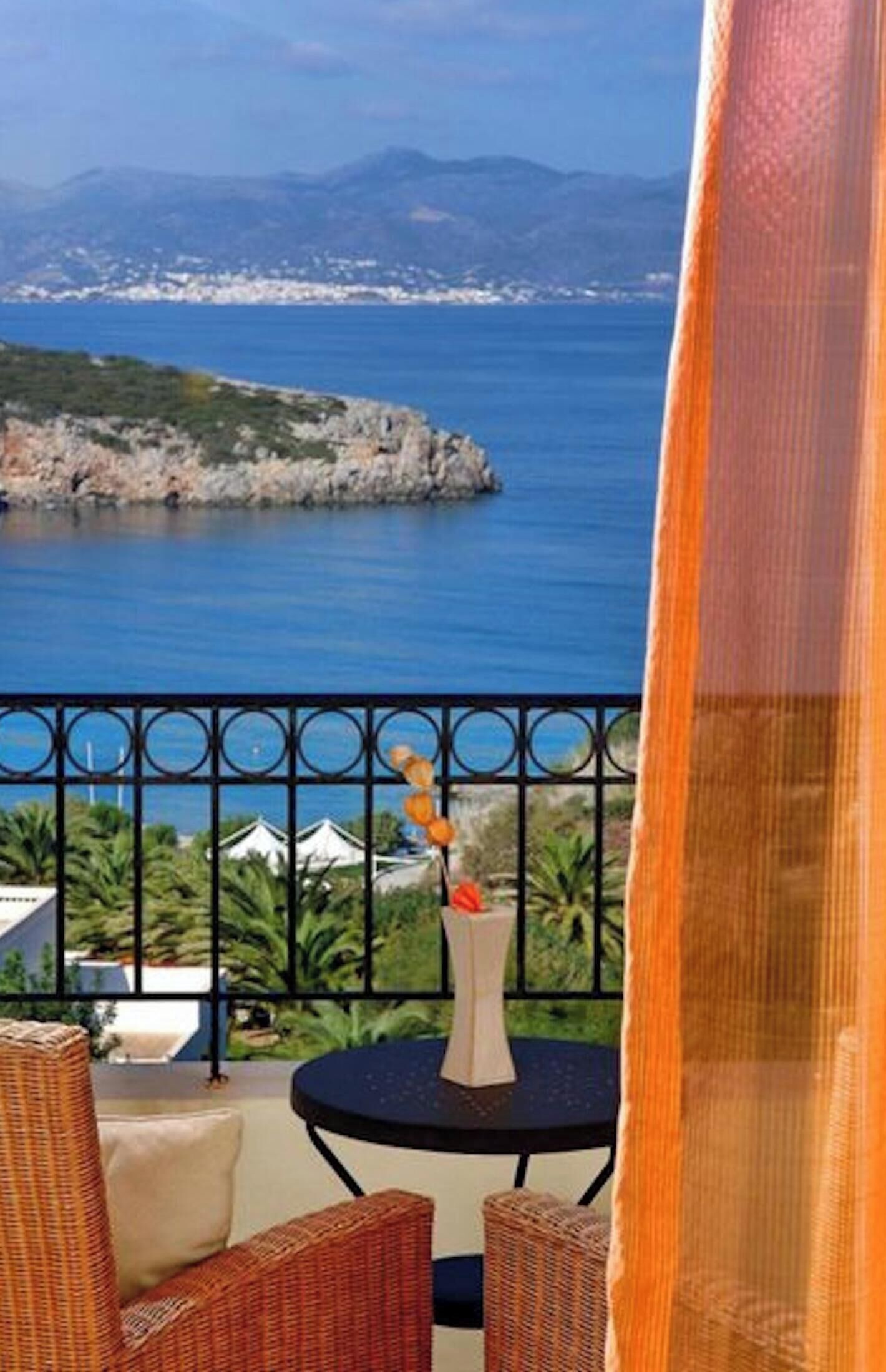 A balcony with wicker furniture, a vase with flowers, and a view of the sea and mountains.