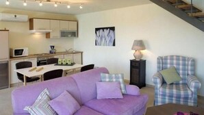 A living room with a purple sofa, a plaid armchair, a dining table with chairs, a kitchen area, and a staircase.