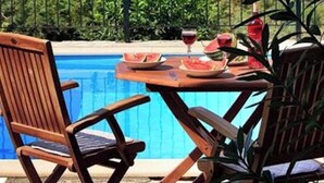 A poolside setting with a view of the sea and mountains, a table with drinks and a watermelon, and two wooden chairs.